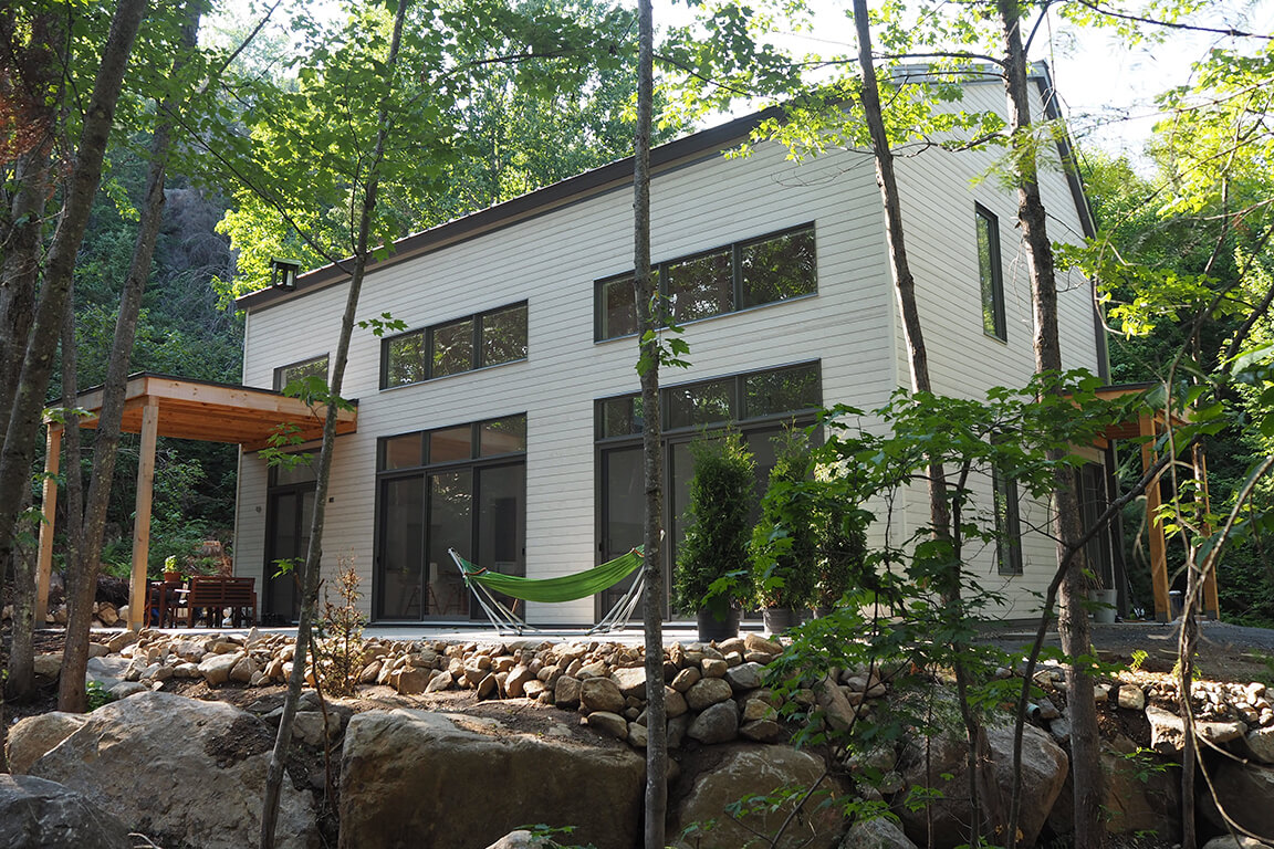 Vue de la façade sud avec sa grande terrasse et sa fenestration abondante dans un environnement boisé, projet La Mésange réalisé par Biophile