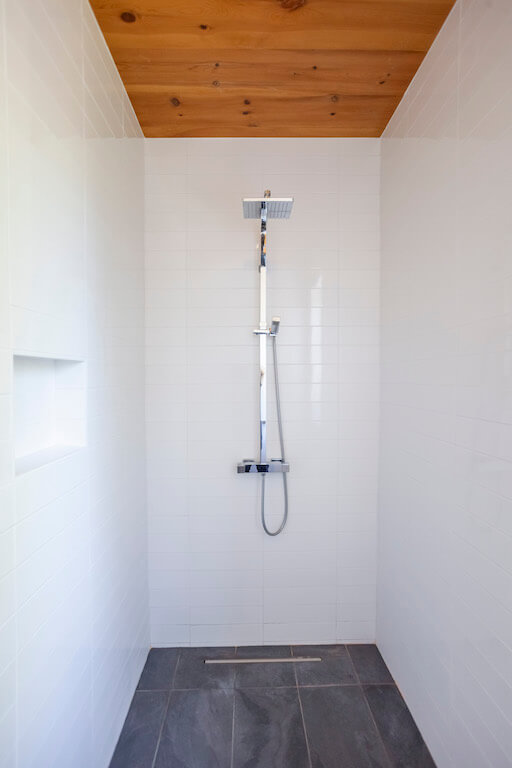 Vue de la douche de la salle de bain au rez-de-chaussée avec plancher d'ardoise et murs recouverts de céramique blanche avec jet de pluie, projet La Grive réalisé par Biophile architecture